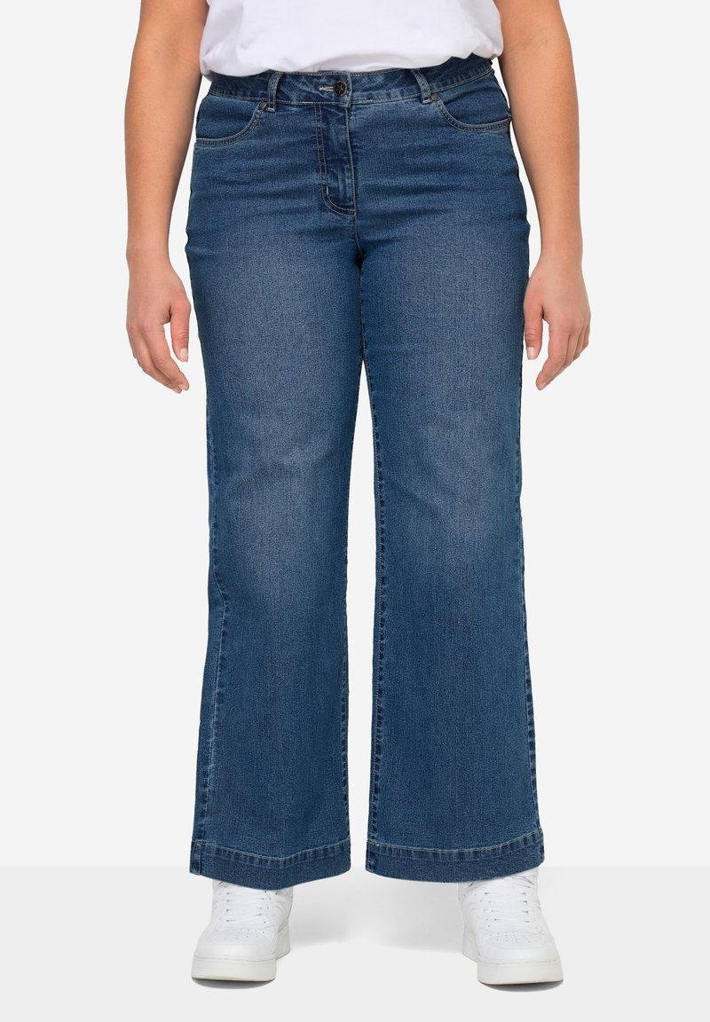 Person trägt mittelhoch geschnittene blaue Jeans mit weitem Bein, vorderen Taschen und weißen Sneakers, steht vor einem schlichten Hintergrund.