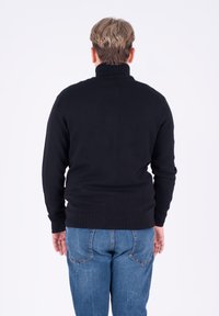Maglione nero a maglia con colletto alto, caratterizzato da polsini e orlo a coste, indossato sopra dei jeans blu, visto da dietro.