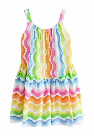 Ragazza giovane con capelli ricci che indossa un abito senza maniche a righe ondulate arcobaleno e sandali bianchi, in piedi davanti a uno sfondo semplice.