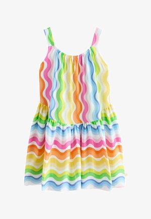 Ung pige med krøllet hår iført en ærmeløs kjole med regnbuefarvede bølgede striber og hvide sandaler, der står mod en ensfarvet baggrund.