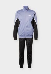 Μοβ και μαύρη φόρμα με ζακέτα με φερμουάρ και ψηλό γιακά, διακοσμημένη με ρίγες, σε συνδυασμό με αντίστοιχο μαύρο παντελόνι. Λογότυπο PUMA και στα δύο κομμάτια.