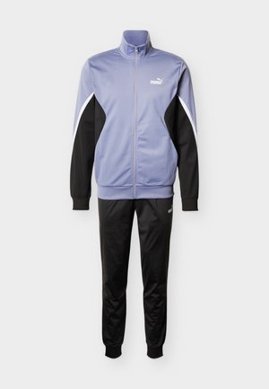 Vijolična in črna trenirka z zadrgo, visokim ovratnikom in črtami, v paru z usklajenimi črnimi hlačami. Na obeh kosih je logo PUMA.