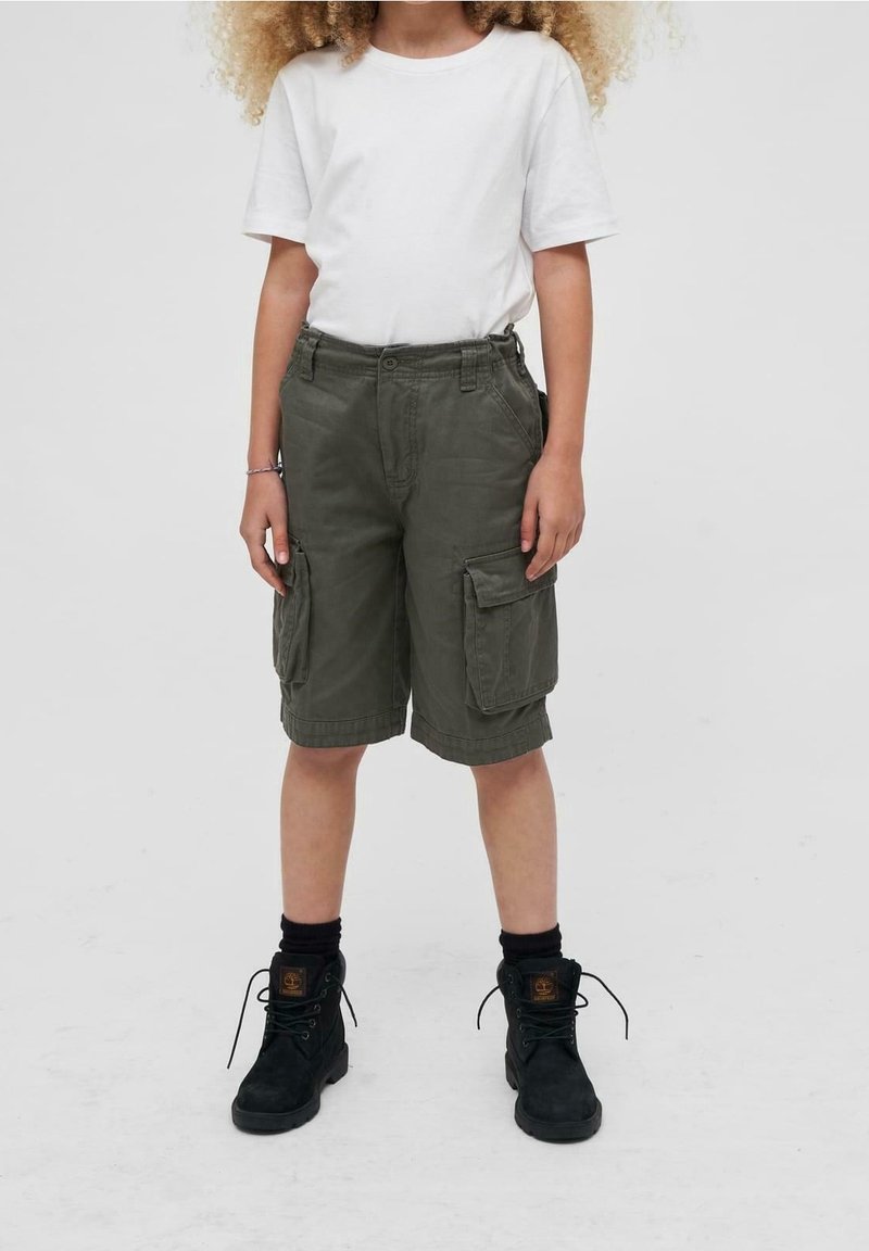 Enfant portant un t-shirt blanc, un short cargo vert foncé, des chaussettes noires et des bottes noires à lacets, debout devant un fond uni.