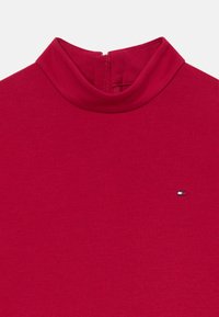 Crvena turtleneck vestica napravljena od glatkog materijala, s kratkim patentnim zatvaračem na leđima i malim logom u donjem lijevom kutu.