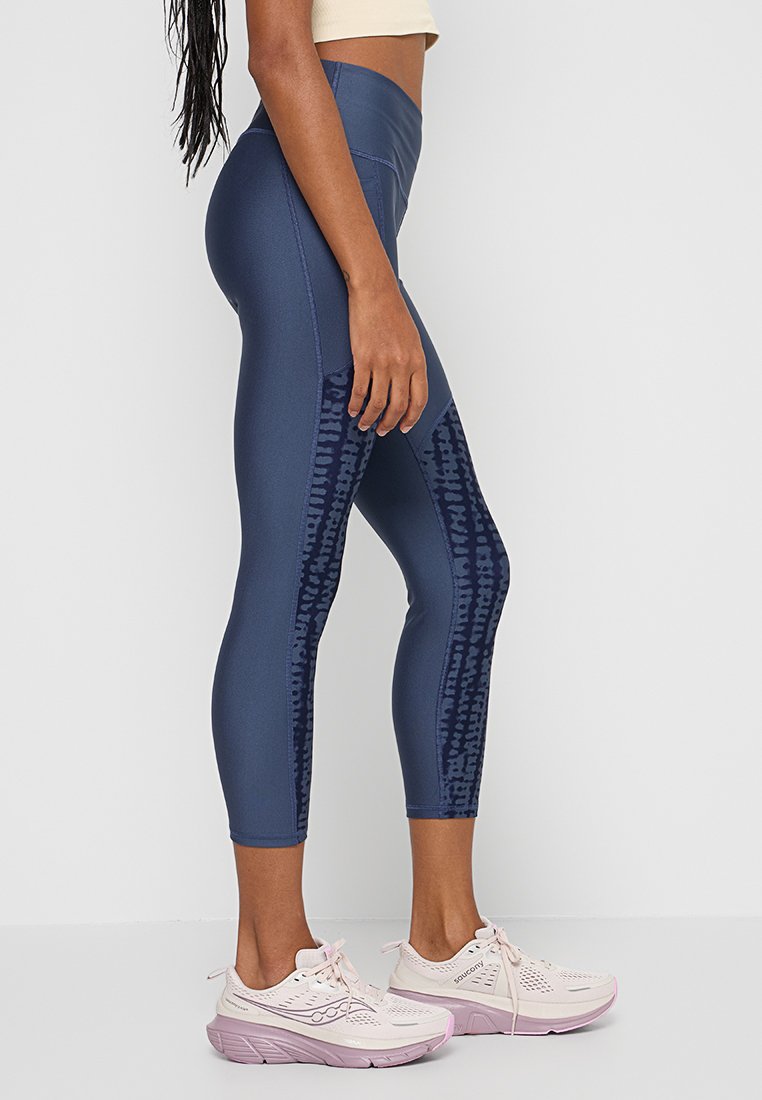 Mujer vestida con leggings de estampado azul marino y zapatillas de correr rosa claro, de pie contra un fondo blanco, vista lateral.