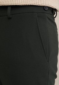Pantaloni personalizați de culoare verde închis, confecționați dintr-un material texturat, având buzunare laterale, bucle pentru curea și un închizător cu buton negru la talie.