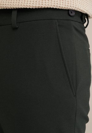 Temno zelene hlače krojenega sloga iz teksturirane tkanine, ki vključujejo stranske žepe, zakovico za pas in črno gumbno zapiranje v pasu.