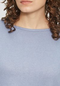 Vrouw met krullend bruin haar, gekleed in een lichtblauwe gebreide top met een ronde halslijn, van kin tot bovenste borst weergegeven.