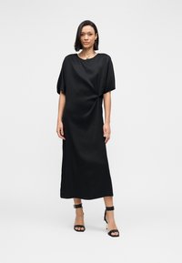 Schwarzes, bodenlanges Kleid mit kurzen, weiten Ärmeln und einem geknoteten Detail an der Taille. Gefertigt aus glattem Stoff mit einer drapierten Silhouette.