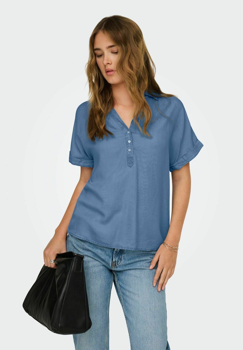 Femme aux cheveux ondulés portant un chemisier bleu à manches courtes boutonné et un jean, tenant un sac à main noir dans sa main gauche.