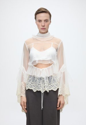 Άτομο με κοντά μαλλιά που φοράει διάφανη λευκή δαντελένια μπλούζα πάνω από λευκό τοπ και σκούρο παντελόνι με φαρδιά μπατζάκια, στέκεται μπροστά σε απλό φόντο.