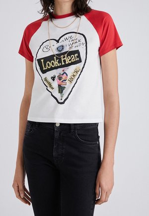 Mujer con camiseta corta blanca de mangas rojas y un gráfico de corazón con el texto "Mira. Escucha." junto con detalles artísticos coloridos.