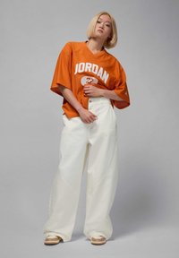 Orange mesh jersey with "JORDAN" logo, paired with white wide-leg pants featuring curved seams and beige shoes with layered design.