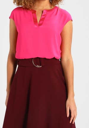 Kobieta ubrana w jasnoróżową bluzkę z krótkim rękawem, wpuszczoną w wysokowoską ciemnorubinową spódnicę z paskiem i okrągłą klamrą.