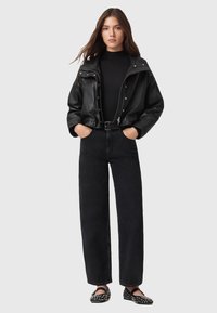 Giacca bomber nera in pelle con bottoni metallici, abbinata a jeans neri a vita alta e ballerine nere con borchie, creando un look monocromatico.
