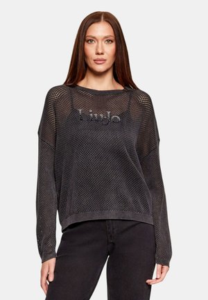 Donna con lunghi capelli castani che indossa un maglione nero in rete sopra una maglietta nera e pantaloni neri, in piedi contro uno sfondo semplice.