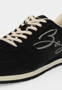 Baskets noires avec lacets beige, bande blanche sur la semelle et grand logo gris en écriture cursive sur le côté.
