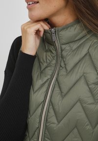 Olivgrünes, gesteppte Weste mit Chevron-Muster, Reißverschluss und schwarzen Rippärmel. Weiches, leichtes Material mit einer glatten Textur.