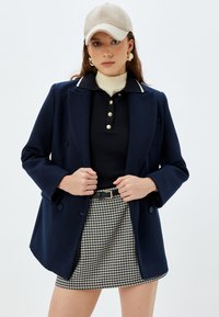 Blazer doppiopetto blu, in tessuto misto lana, con bordi bianchi al colletto. Polo nera con bottoni dorati. Gonna mini a scacchiera, cintura nera.