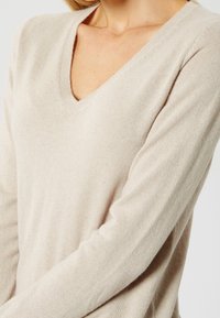 Femme portant un pull beige à manches longues avec un col en V, visible des épaules à la taille, sur un fond uni.