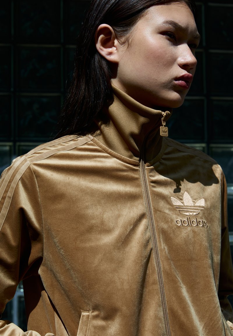 Braune Samt-Zip-Jacke mit hohem Kragen, Seitentaschen und gesticktem Logo. Drei Streifen-Details an den Ärmeln, glatte Textur.