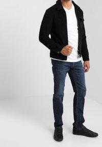 Chaqueta de pana negra con cuello, cierre de cremallera y dos bolsillos frontales, combinada con una camiseta blanca y jeans azules, finalizada con zapatillas negras.