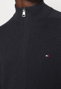 Maglione grigio scuro lavorato a maglia con colletto alto e zip argentata; presenta un piccolo logo in rosso, bianco e blu. Texture liscia.