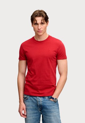 Joven con cabello castaño ondulado vistiendo una camiseta roja lisa de manga corta, vaqueros azules y un reloj de pulsera con correa azul.