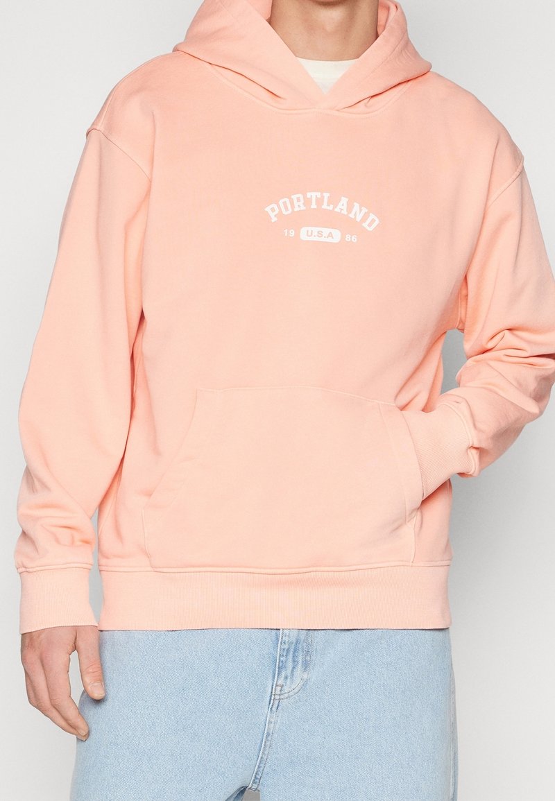 Pfirsichfarbener Hoodie aus weichem Material, ausgestattet mit einer Kängurutasche, einer Zugkordelkapuze und weißen Aufdrucktexten "PORTLAND 1986."