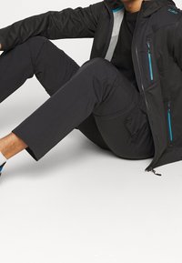 Veste noire avec accents turquoise, poches zippées et capuche, associée à un pantalon foncé. Texture lisse, design moderne, coupe athlétique.