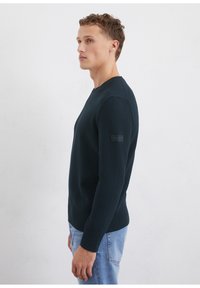 Marineblaue, gerippte Strickjacke mit Rundhalsausschnitt, slim fit und einem kleinen Logopatch am Ärmel. Weiche Textur und lange Ärmel.