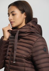 Braune Pufferjacke mit hohem Kragen, gestepptem Design und verstellbarem Kordelzug. Auffälliger schwarzer Logo-Patch auf der linken Schulter.
