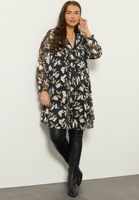 Zwarte bloemen tuniekjurk met lange doorschijnende mouwen en een v-hals, gecombineerd met zwarte leggings en enkellaarsjes. Heeft een losse pasvorm en een fijn patroon.