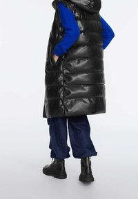 Schwarze Steppweste mit Kapuze, Reißverschlusstaschen und gestepptem Design. Getragen über einem blauen Langarmhemd und kombiniert mit blauen Jeans und schwarzen Stiefeln.