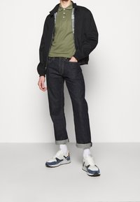 Svart bomberjacka över en grön polo skjorta, kombinerad med mörka denimjeans och vita sneakers med blå och orange detaljer.