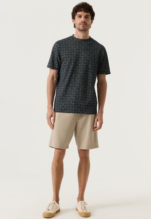 Man die staat, draagt een donker T-shirt met patroon en korte mouwen, beige knielange shorts en beige veter-sneakers op een witte achtergrond.