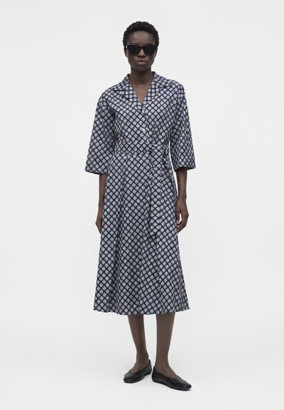 Γυναίκα με μπλε σκούρο φόρεμα με σχέδια και 3/4 μανίκια, μαύρες μπαλαρίνες και μαύρα γυαλιά ηλίου, στέκεται μπροστά σε απλό λευκό φόντο.