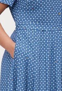 Mėlyna suknelė su balta ir šviesiai mėlyna deimanto raštu, su klostėmis ir šoninėmis kišenėmis. Medžiaga yra glotni su struktūrizuotu dizainu.