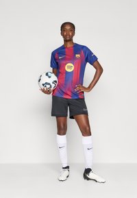 FC Barcelona női labdarúgó mez, kék és piros csíkos felsővel, fekete rövidnadrággal, fehér zoknival, és egy labdát tartva. Nike logó látható.