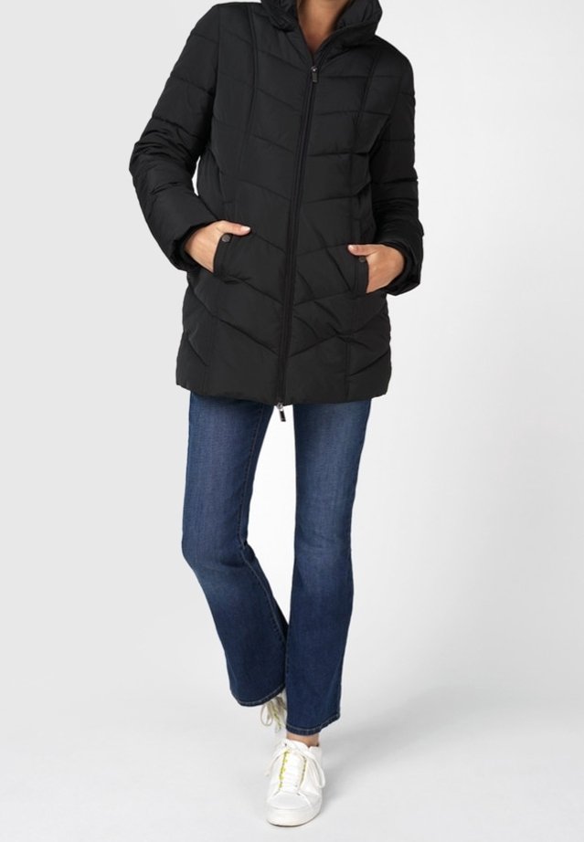 Femme portant une doudoune noire zippée, un jean bleu et des baskets blanches, debout avec les mains dans les poches de la veste.