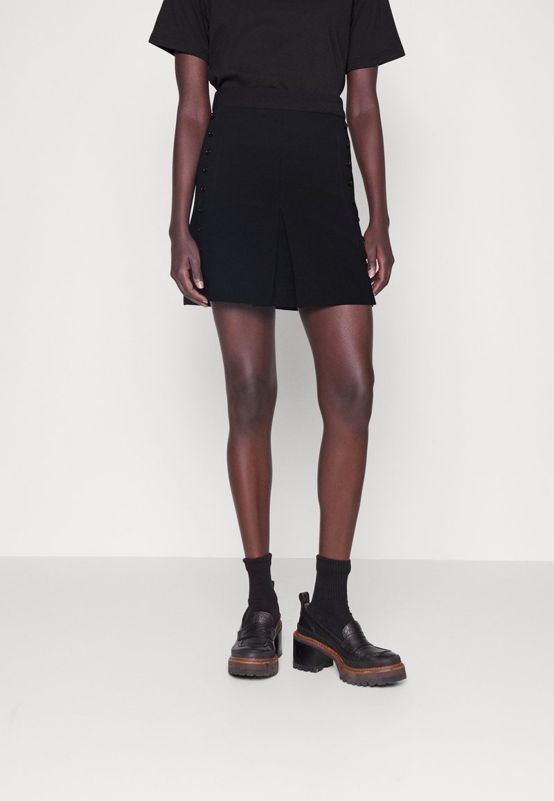 Personne portant un haut noir à manches courtes, une mini-jupe noire avec des détails de boutons sur le côté, des chaussettes noires et des chaussures plateformes noires épaisses.