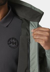 Donkergrijze polo van textiel met een zwarte logo, onder een groene vest met rits, voorzien van een zachte fleece voering en contrasterende panelen.