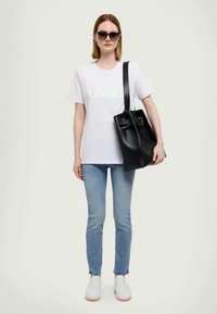 Camiseta branca com logótipo, jeans de tom azul claro, sapatilhas brancas e uma bolsa tote preta com fivela. Modelo a usar óculos de sol escuros.