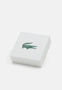 Boîte rectangulaire blanche avec une surface texturée, arborant un logo de crocodile vert sur le dessus. Design fermé sans quincaillerie visible.