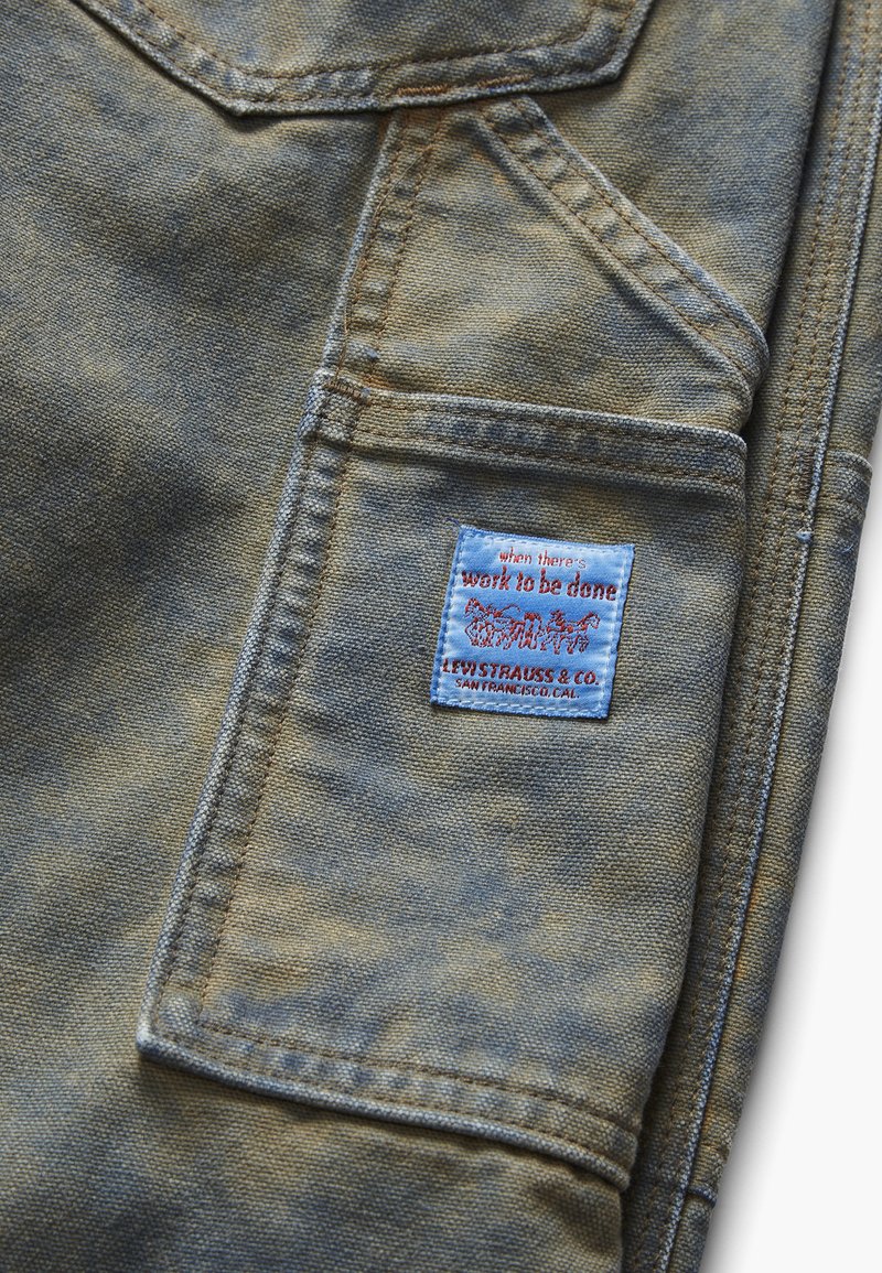 Detail vyblednutého džínového vrecka s modrým štítkom Levi Strauss & Co., na ktorom je nápis "when there's work to be done" a logo dvoch koní ťahajúcich džínsy.