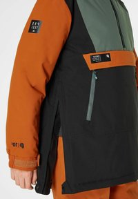 Isolierte Jacke mit schwarzen, orangen und grauen Einsätzen, Reißverschlusstaschen, verstellbaren Ärmelbündchen und einem Markenpatch auf dem Ärmel.