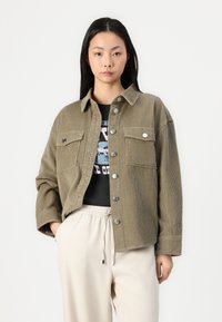 Veste en velours côtelé vert olive avec des boutons argentés, deux poches poitrine et une coupe décontractée, portée sur un t-shirt noir à motif et un pantalon clair.