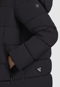 Schwarze Steppjacke mit einem Reißverschlussfach, Logo-Patch und einem gesteppten Design mit matter Oberfläche. Der Ärmel hat einen gerippten Bund.