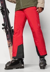 Pantalons de ski rouges avec des accents noirs, fabriqués en tissu durable. Caractéristiques : coupe décontractée et poches inférieures renforcées, accompagnés de bottes de ski noires et de skis.