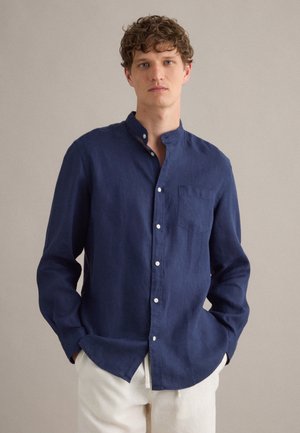Giovane uomo con capelli ricci che indossa una camicia blu navy con collo alla mandarino abbottonata e pantaloni bianchi, in piedi con le mani nelle tasche.